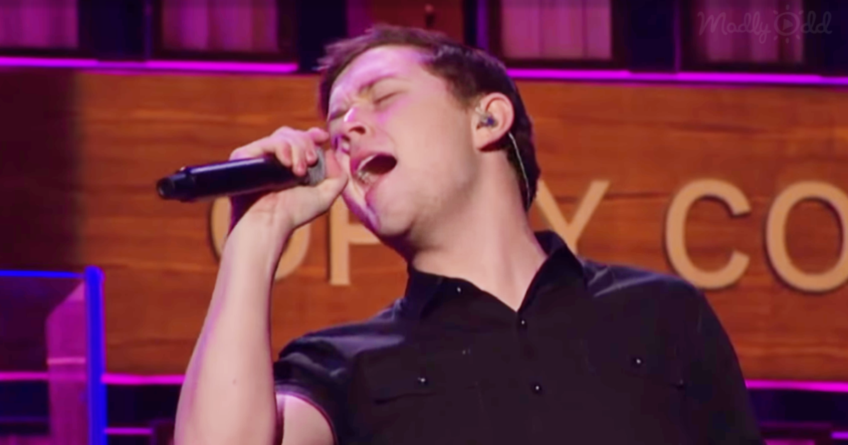 A person in a black shirt sings passionately into a microphone on a stage with purple lighting.