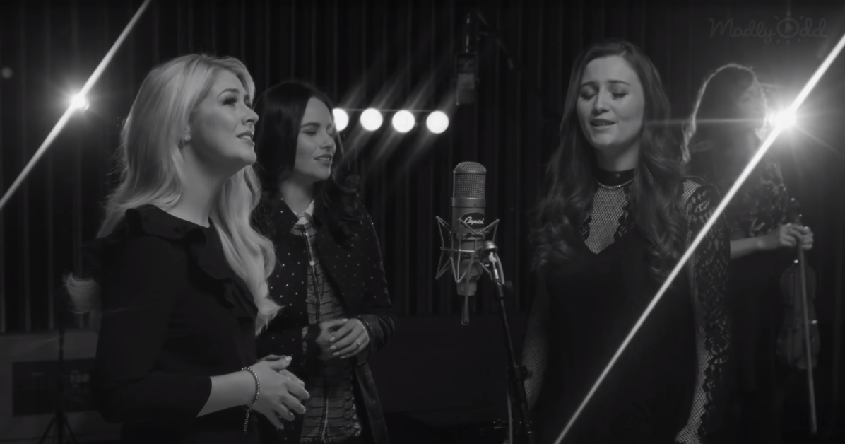 Three women sing in a dimly lit studio with a microphone and lights.
