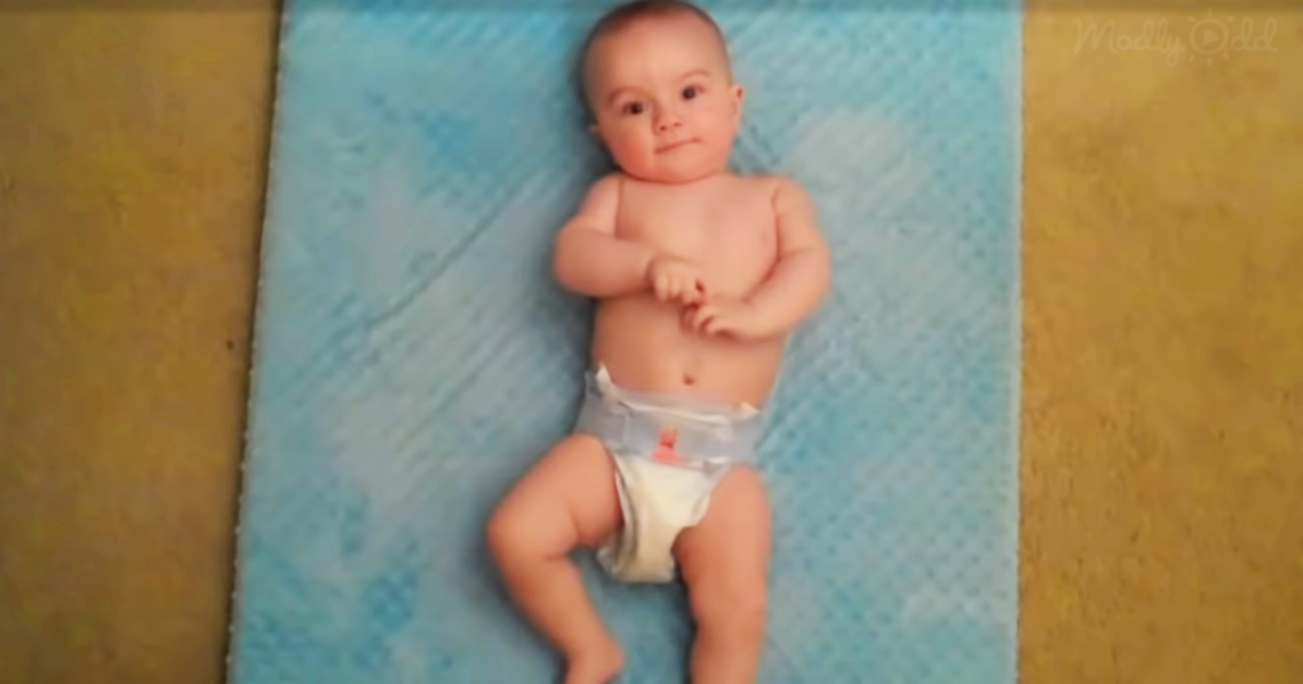 A baby lies on a blue mat, wearing a diaper, with arms crossed and a curious expression.
