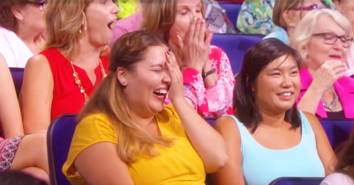 A woman in a yellow shirt laughs heartily, covering her face with her hand, surrounded by other audience members.