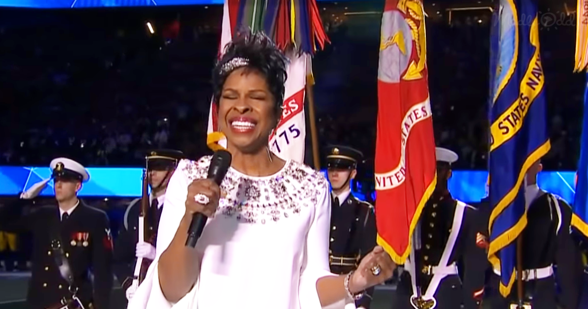 A woman in white attire sings into a microphone, surrounded by military personnel and flags.