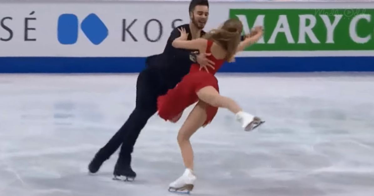 A figure skating couple performs a dynamic lift on ice, with the man supporting the woman in a dramatic pose.