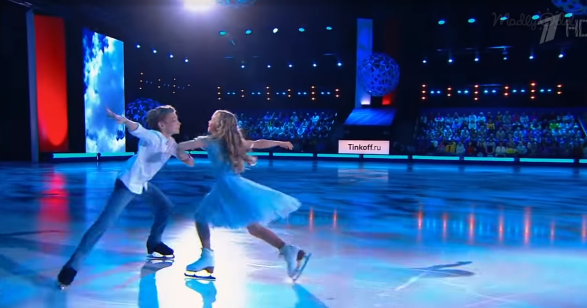 A boy and girl in ice skating attire perform a synchronized move on an ice rink.