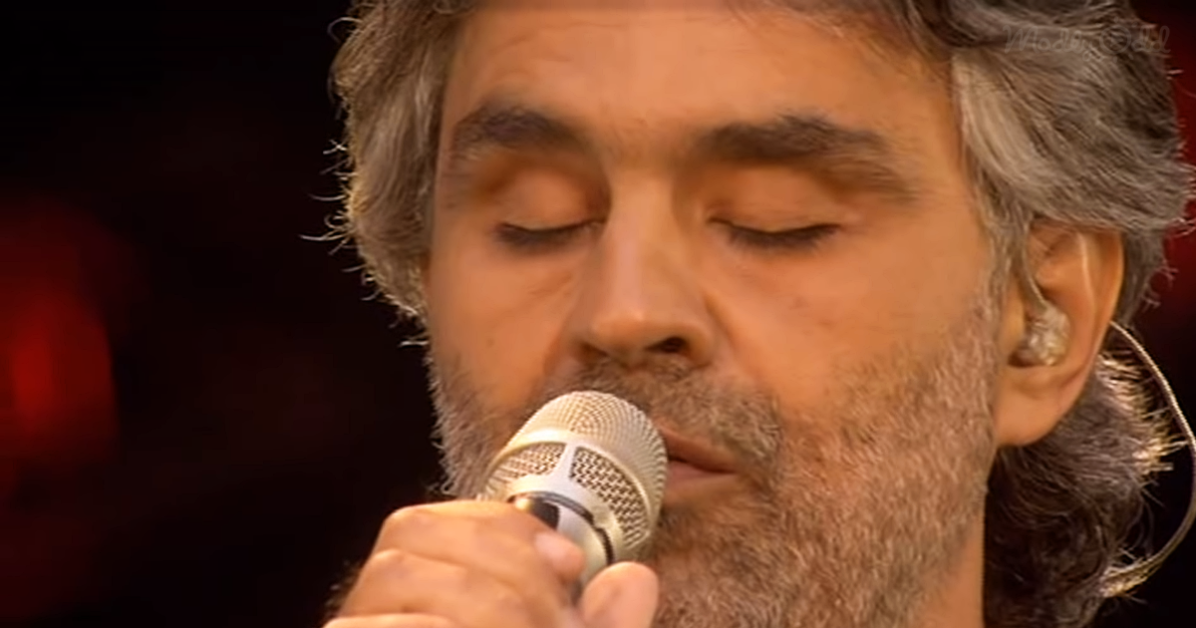 A man with gray hair and beard sings into a microphone with his eyes closed.