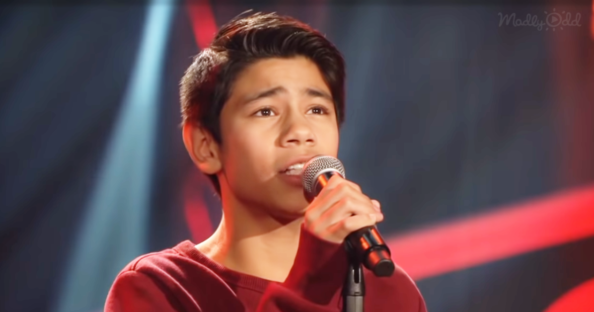 A young person in a red shirt sings into a microphone on a stage with red and blue lighting.