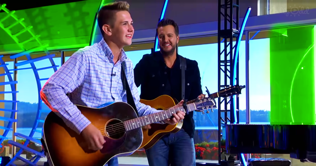 A man in a plaid shirt plays an acoustic guitar on a stage with another man in a black jacket standing behind him.