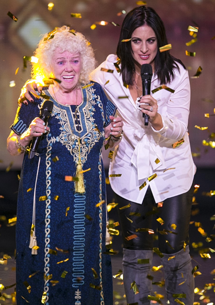 Two women, one in traditional attire and the other in white, hold microphones amidst falling confetti, celebrating on stage.