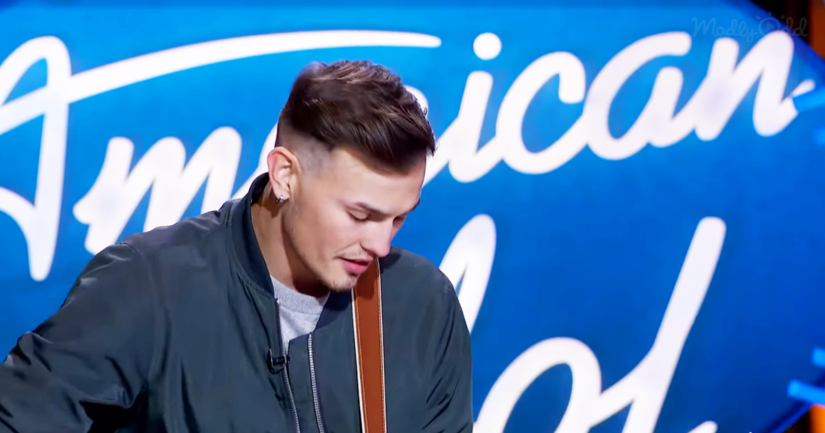 A man in a dark jacket stands in front of a blue "American Idol" backdrop, looking down.