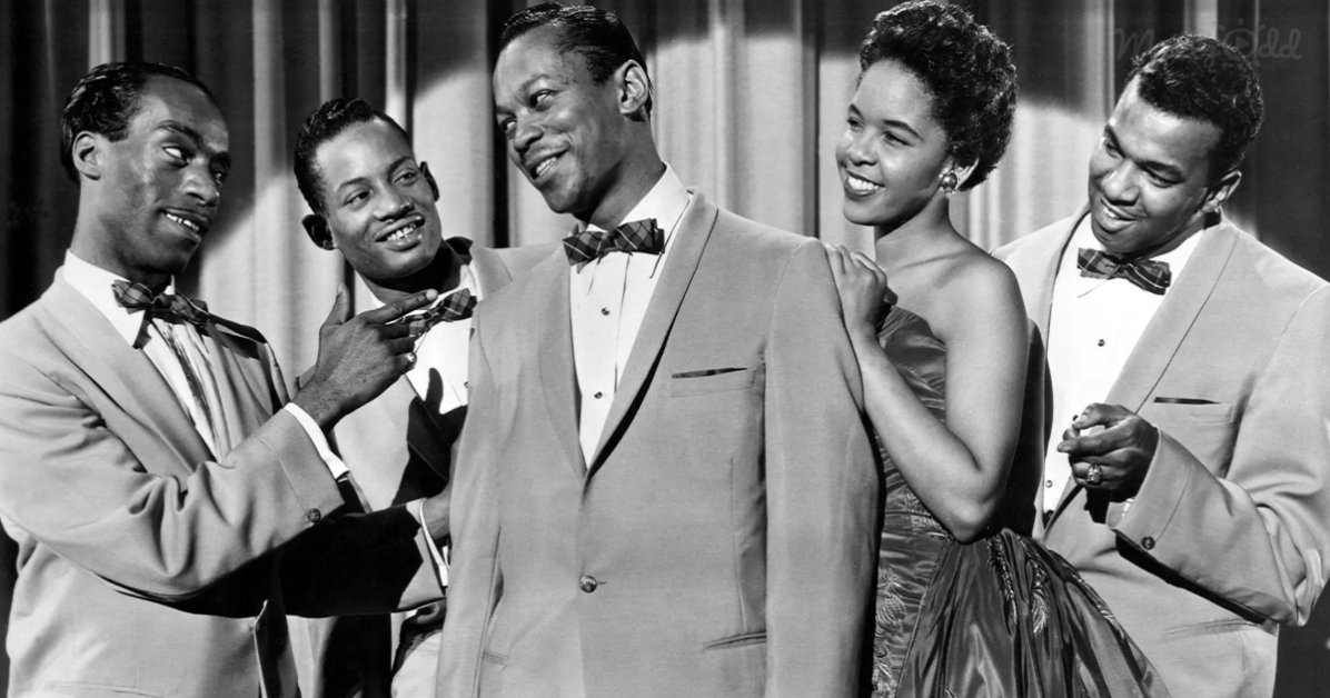 A man in a suit and bow tie is surrounded by smiling people in formal attire, suggesting a celebratory or formal event.