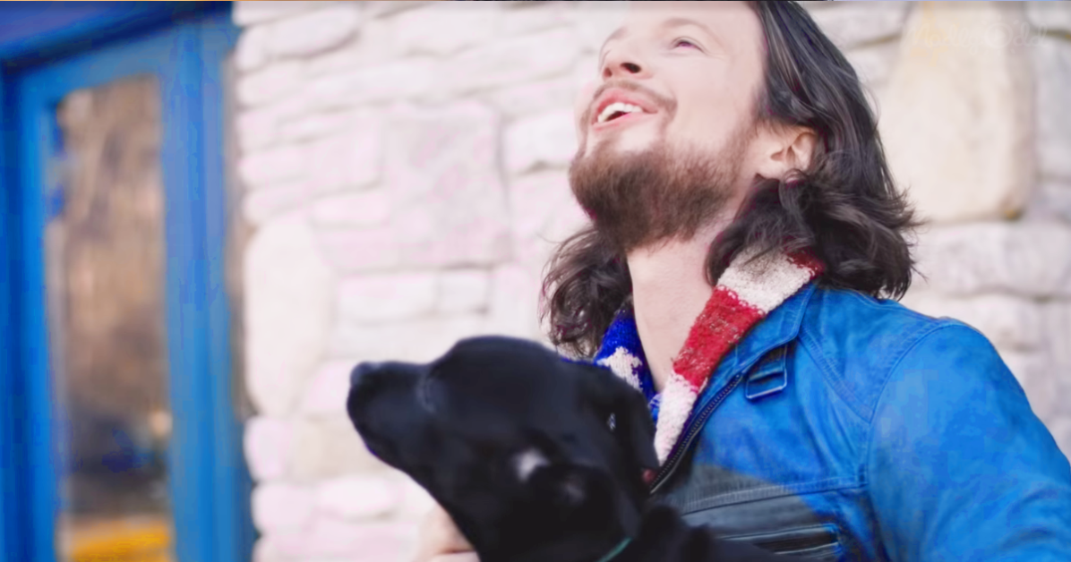 A man with long hair and a beard, wearing a blue jacket, looks up smiling while holding a black dog.