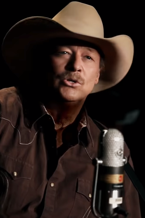 A man in a cowboy hat and brown shirt sings into a microphone against a dark background.