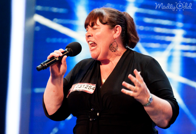 A woman in a black outfit sings passionately into a microphone on a stage with blue lighting.