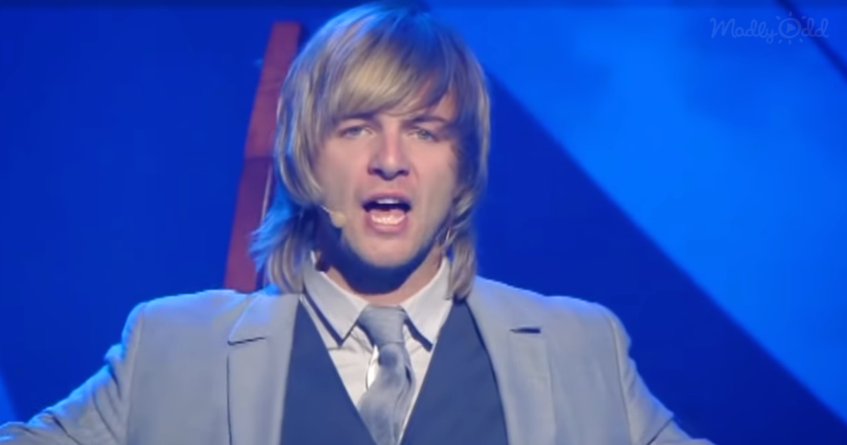 A man with long blonde hair, wearing a light gray suit and tie, is singing on stage with a microphone.