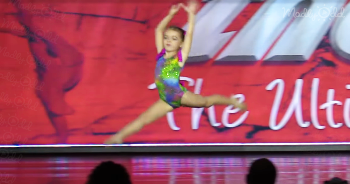 A young gymnast in a colorful leotard performs a split leap on a stage with a red background.