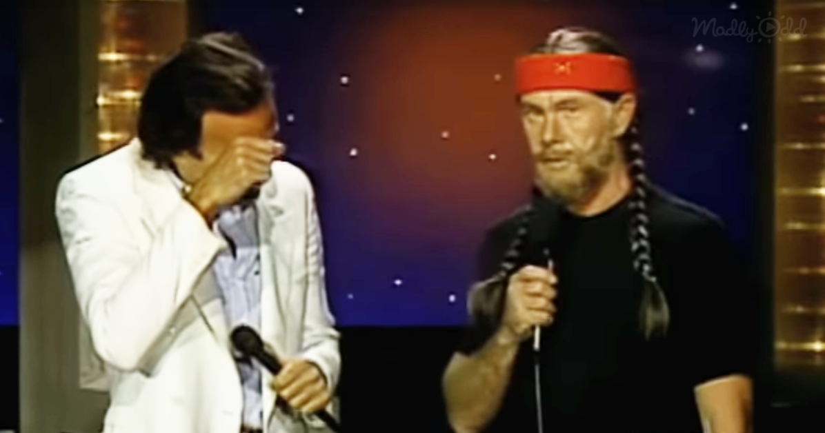 A man in a white suit holds a microphone, while another man in a black shirt and red headband speaks into a microphone.