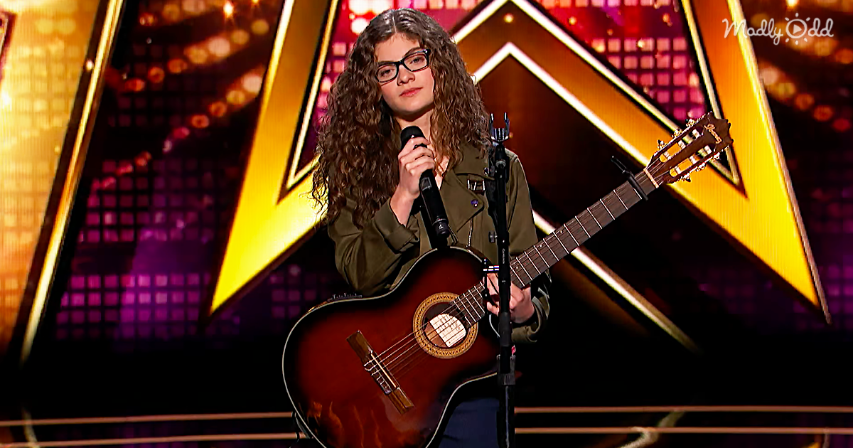 A person with curly hair and glasses holds a microphone and guitar on stage with a colorful background.
