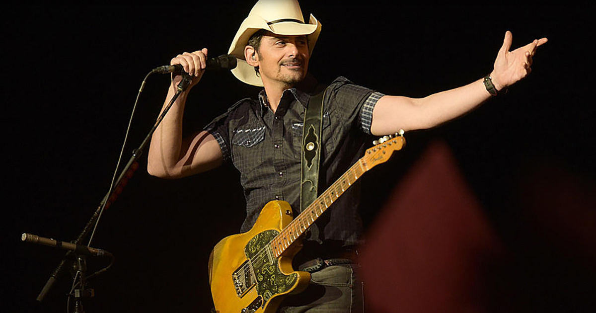 Brad Paisley performs on stage, holding a guitar and pointing, wearing a cowboy hat and plaid shirt.