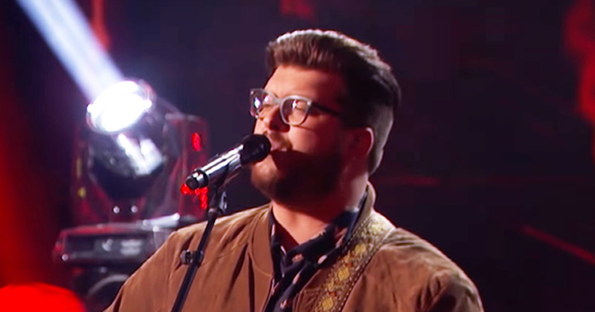 A man in glasses plays guitar and sings into a microphone, with a spotlight and red circular logo in the background.