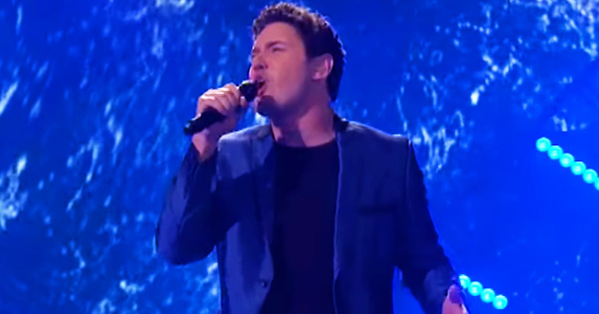 A singer in a dark suit performs on stage, holding a microphone, with a blue background and text promoting "Somewhere"...