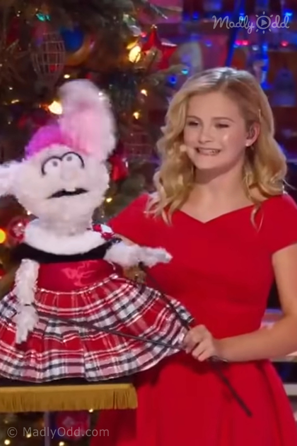 A young woman in a red dress holds a puppet dressed in a plaid outfit, with a festive Christmas tree in the background.