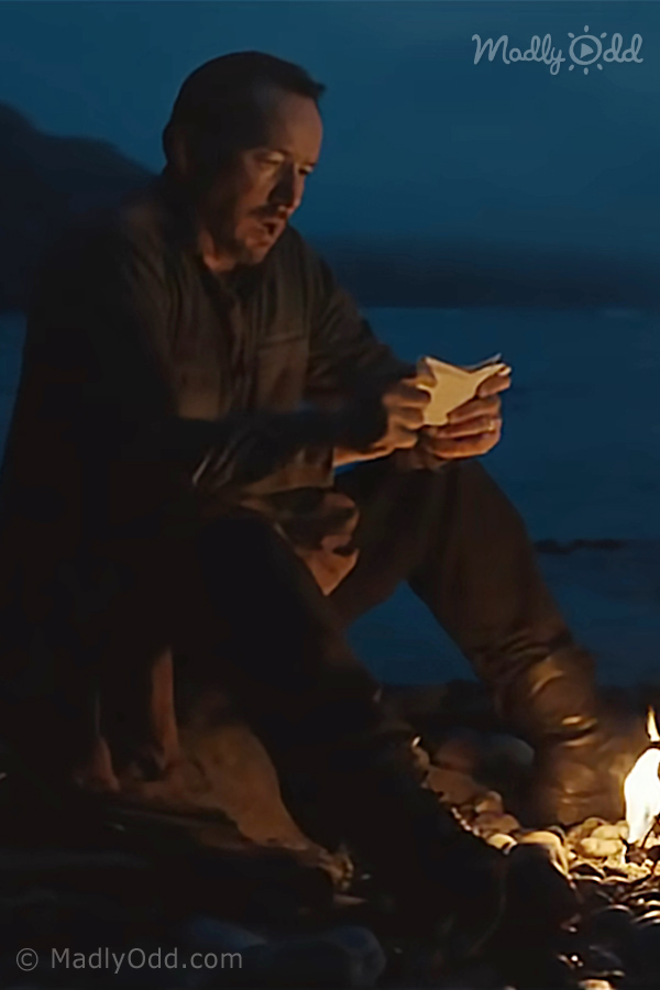 A man sits by a campfire, holding a phone, with a dark, serene background.
