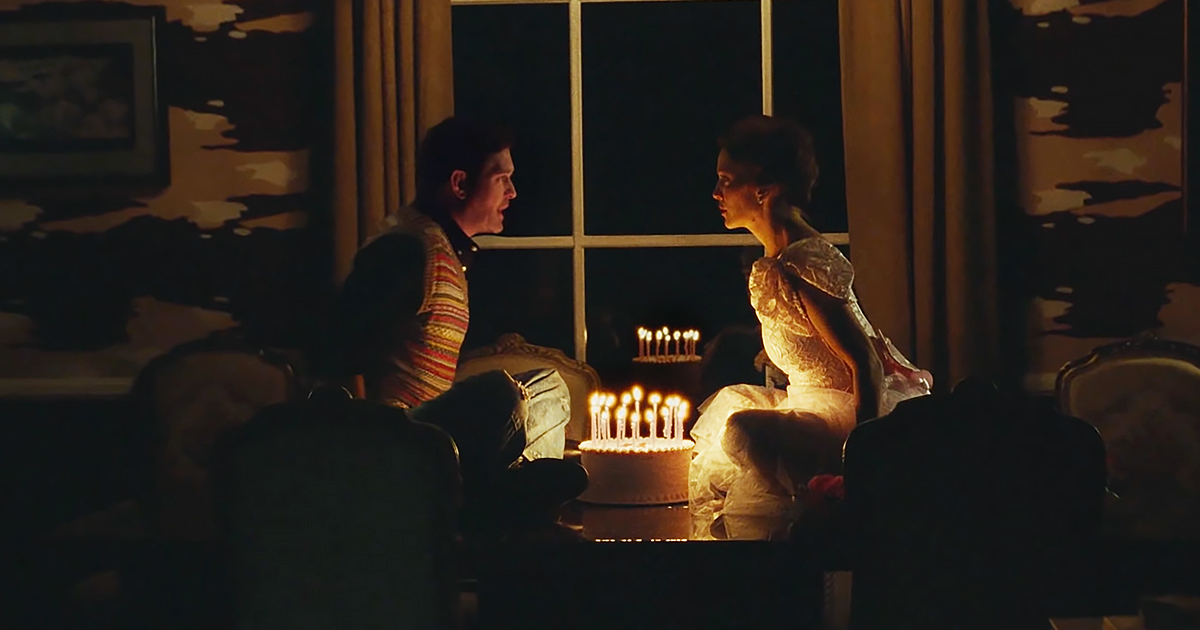 A couple sits by a lit birthday cake, surrounded by dimly lit room with a window and curtains.