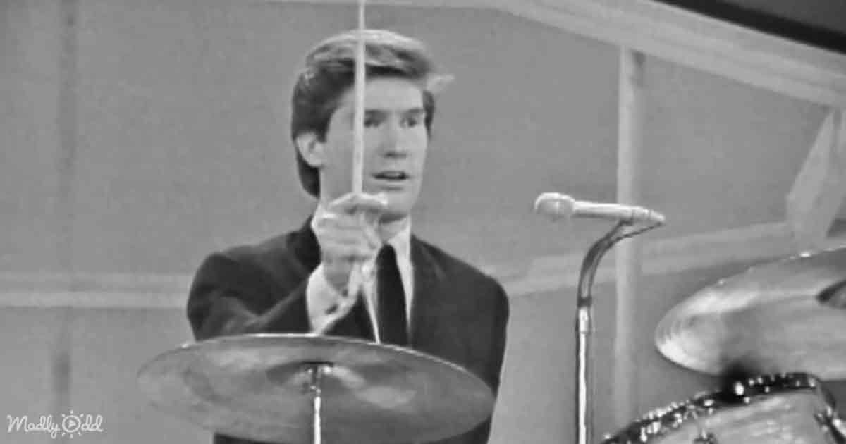 A man in a suit plays drums, holding a drumstick, with a microphone and cymbals visible.