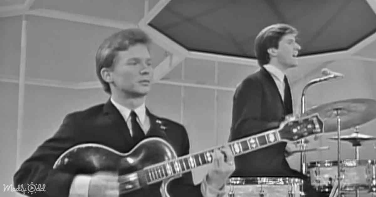 A man in a suit plays guitar while another sings and plays drums in a recording studio.