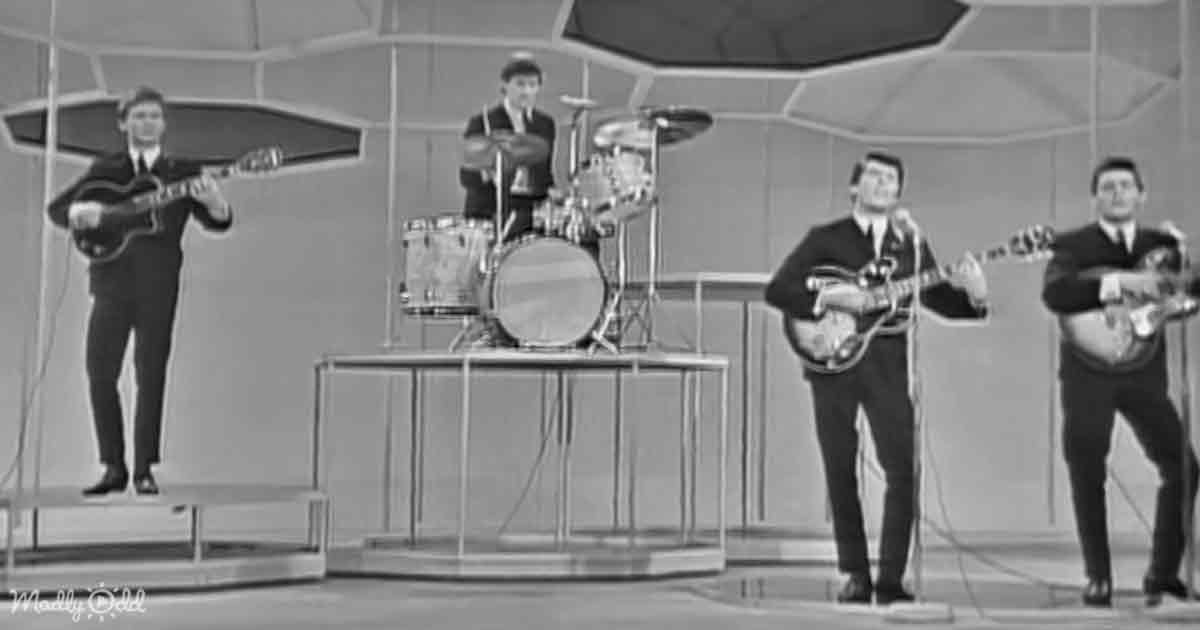 A band performs on stage with a drummer, two guitarists, and a bassist, all dressed in suits.