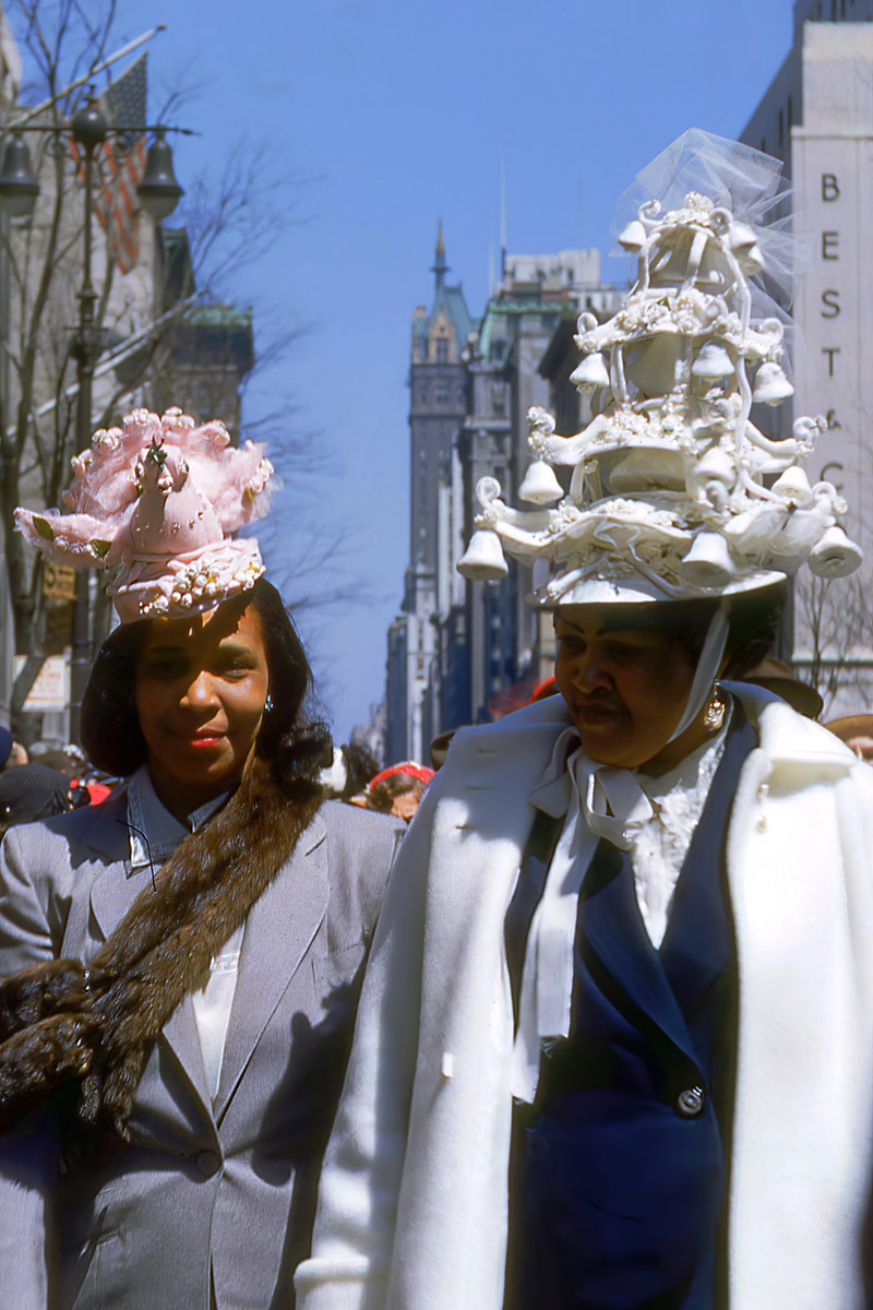 Two women in elaborate hats, one pink, one white, stand on a city street with a tall building in the background.