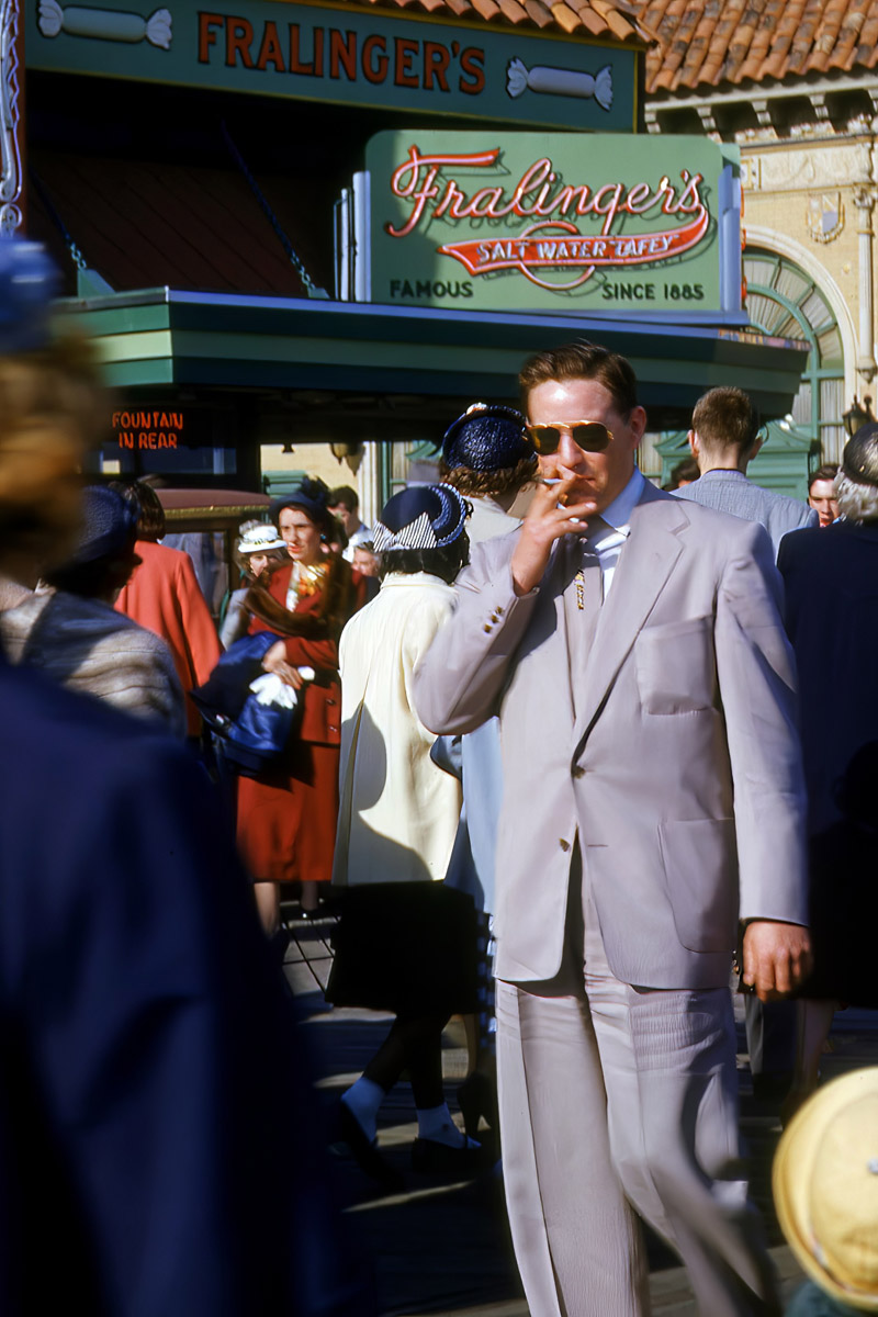 A man in a light suit smokes a cigarette, wearing sunglasses, amidst a crowd near Fralinger's Salt Water Taffy stand.