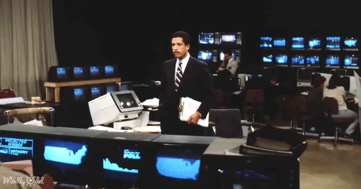 A man in a suit stands in a dimly lit room filled with monitors and computers, holding papers.